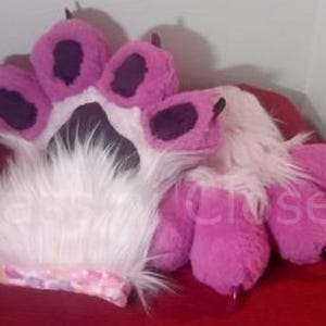 May include: A pair of pink and white furry paw hand puppets with black claws. The puppets are made of soft, plush fabric and have a realistic paw shape.