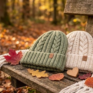 Puede incluir: Dos gorros de punto, uno verde salvia y otro crema, con patr&oacute;n de trenzas y borde doblado. Cada gorro tiene una peque&ntilde;a etiqueta cuadrada marr&oacute;n con un icono de coraz&oacute;n en la parte frontal. Una bufanda de punto crema est&aacute; colocada debajo.