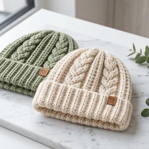 Puede incluir: Dos gorros con vuelta, uno en verde salvia y otro en crema, con un patr&oacute;n de punto de ochos grueso y un borde acanalado. Cada gorro incluye una peque&ntilde;a etiqueta cuadrada de piel sint&eacute;tica color canela en el borde frontal con un coraz&oacute;n.