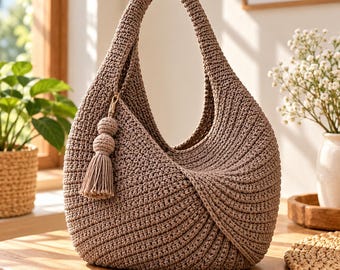 May include: A taupe-colored hobo-style handbag crafted from thick, textured crochet yarn. The bag features a single integrated handle, a rounded silhouette, and a decorative double-sphere tassel accent attached to the side with a gold-toned clasp.