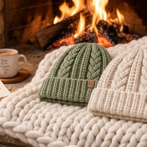 Puede incluir: Dos gorros de punto con vuelta, uno verde salvia y otro crema, con un patr&oacute;n de trenzas verticales y una peque&ntilde;a etiqueta cuadrada marr&oacute;n con un icono de coraz&oacute;n en la vuelta. Los gorros descansan sobre una manta blanca de punto grueso.