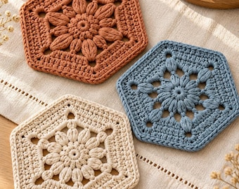 May include: Three hexagonal crochet coasters in terracotta, dusty blue, and cream. Each coaster features a central floral motif with puff stitches and an openwork border, showcasing intricate yarn work and a textured finish.