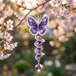 May include: A purple and white crocheted butterfly ornament hanging from a tree branch. The butterfly features intricate stitching, small white floral accents, and a spiral tail ending in a faceted purple crystal drop suspended by a thin white string.