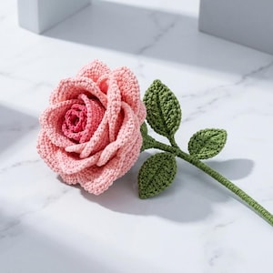 May include: A handcrafted crochet rose featuring layered petals in shades of light pink and a darker pink center. The flower is attached to a green crocheted stem with three detailed green leaves extending from the side, showcasing intricate yarn texture.