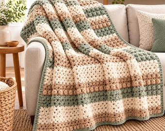 May include: A crochet throw blanket featuring horizontal stripes in sage green, cream, and tan. The blanket is constructed with a textured bobble stitch pattern, creating a raised, rounded surface throughout the fabric design.