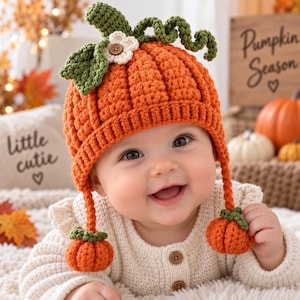 Puede incluir: Un gorro de ganchillo naranja brillante con forma de calabaza, textura acanalada, tallo verde, zarcillos rizados y una pequeña flor blanca con centro marrón. Dos largas orejeras trenzadas cuelgan, cada una con un pequeño adorno de calabaza.