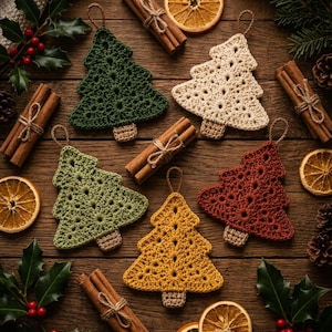 May include: Five crocheted Christmas tree ornaments in dark green, cream, light green, mustard yellow, and rust red. Each tree features a small brown trunk and a hanging loop, arranged among cinnamon sticks, dried orange slices, and holly.