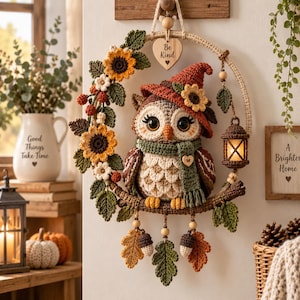Op de afbeelding: Een gehaakte uil wandhanger met een bruine en crèmekleurige uil met een roestkleurige hoed en groene sjaal, zittend op een tak in een cirkelvormig frame versierd met zonnebloemen, bladeren en een lantaarntje, met een hartje met de tekst Be Kind.