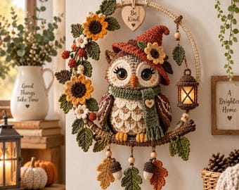 May include: A crochet owl wall hanging featuring a brown and cream owl wearing a rust-colored hat and green scarf, perched on a branch within a circular frame decorated with sunflowers, leaves, and a small lantern, with a heart tag reading Be Kind.