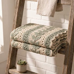 May include: A folded crochet blanket featuring alternating vertical stripes of sage green and cream yarn. The design includes a textured leaf pattern in the green sections and a delicate openwork stitch in the cream sections, finished with a scalloped edge.