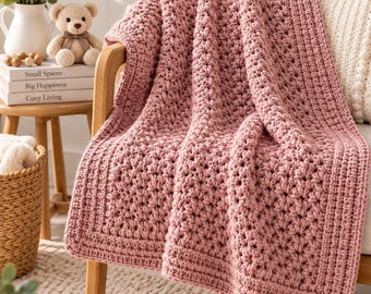 May include: A dusty rose crochet blanket featuring a textured bobble stitch pattern and a solid border. The blanket is draped over a chair, showcasing the intricate yarn work and the soft, muted pink color of the handmade textile accessory.