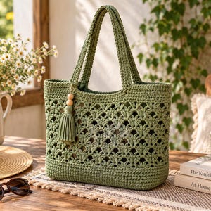 May include: A sage green crochet tote bag featuring an intricate openwork leaf pattern, two sturdy handles, and a decorative tassel accent with two wooden beads attached to one side. The bag has a solid base and a structured rectangular shape.