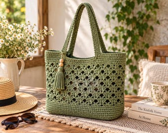 May include: A sage green crochet tote bag featuring an intricate openwork leaf pattern, two sturdy handles, and a decorative tassel accent with two wooden beads attached to one side. The bag has a solid base and a structured rectangular shape.
