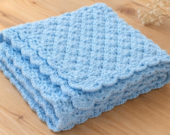 May include: A light blue crochet blanket featuring a textured shell stitch pattern throughout the body and a decorative scalloped edge border. The square-shaped textile is folded into multiple layers, showcasing the intricate yarn work and design.