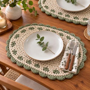 May include: An oval crochet placemat featuring a cream-colored center with a scalloped sage green border. A white ceramic plate sits in the center, topped with a small eucalyptus sprig. A folded napkin with a leaf print, a fork, and a knife are placed on the right side.
