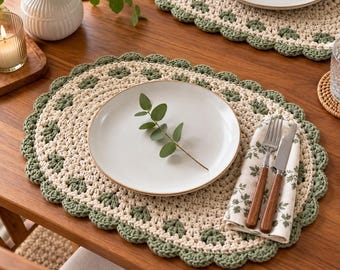 May include: An oval crochet placemat featuring a cream-colored center with a scalloped sage green border. A white ceramic plate sits in the center, topped with a small eucalyptus sprig. A folded napkin with a leaf print, a fork, and a knife are placed on the right side.