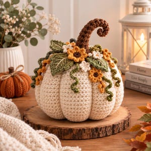 Puede incluir: Una calabaza de ganchillo color crema con un tallo rizado marrón oscuro, adornada con hojas verdes, pequeñas flores blancas y girasoles amarillo dorado tejidos. La calabaza presenta una textura de costillas verticales sobre una base de madera.