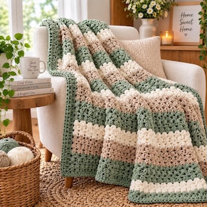 May include: A crocheted throw blanket featuring horizontal stripes in sage green, cream, and beige. The blanket has a textured, open-stitch pattern and is draped over a light-colored armchair. The design consists of repeating color blocks.