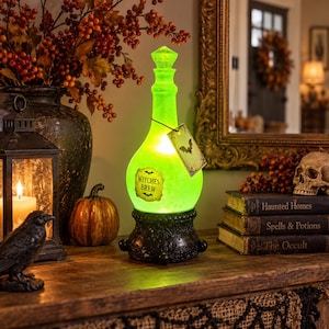 May include: A glowing green glass bottle lamp with a textured finish, resting on a black ornate base. The bottle features a label that reads Witches Brew with a bat illustration and a matching rectangular tag hanging from the neck.