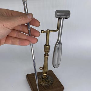 May include: A collection of vintage medical instruments. One instrument is held by a hand. The instruments include a silver-colored tongue depressor with the word "FREEMAN" and a metal hammer and spoon-shaped tool on a brass stand with a wooden base.