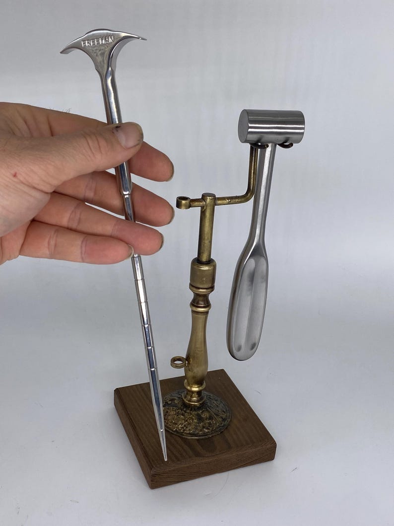May include: A collection of vintage medical instruments. One instrument is held by a hand. The instruments include a silver-colored tongue depressor with the word "FREEMAN" and a metal hammer and spoon-shaped tool on a brass stand with a wooden base.