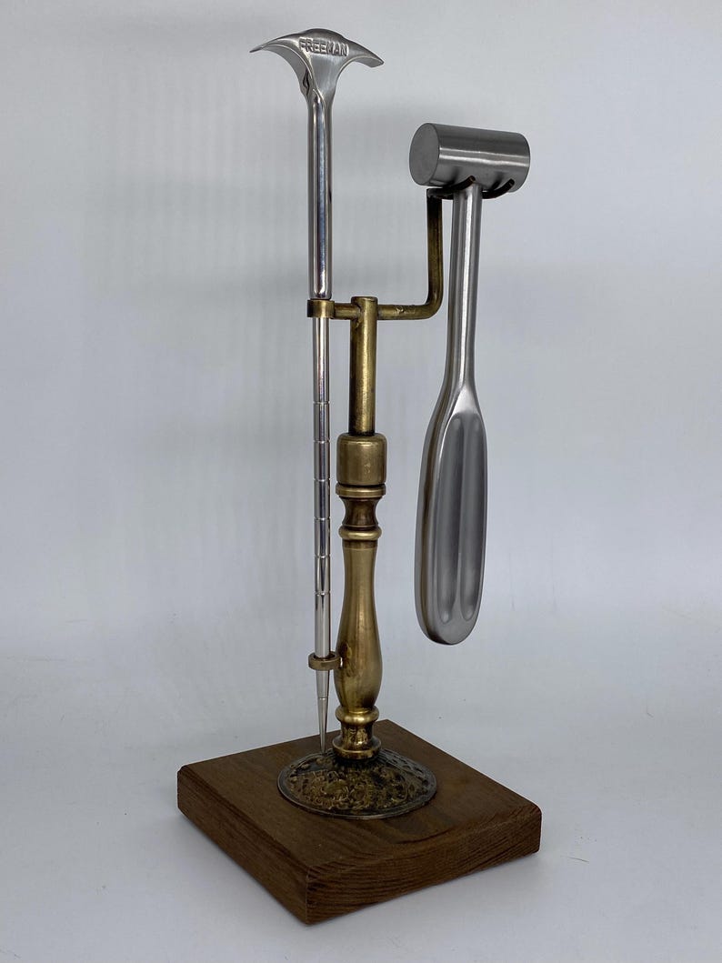 May include: A metal tool set with a wooden base. The set includes a long, slender tool with the word "FREEMAN" on the top, a hammer-like tool, and a spoon-shaped tool. The tools are made of silver and gold-colored metal.