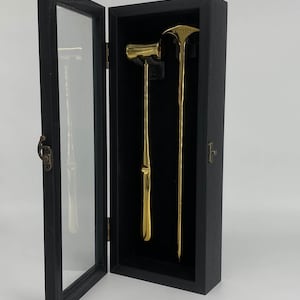 May include: A black wooden display case with a glass door, open to reveal two gold-colored tools. One tool has a hammer-like head, and the other is a long, slender tool with a pointed end. The case is set against a white background.