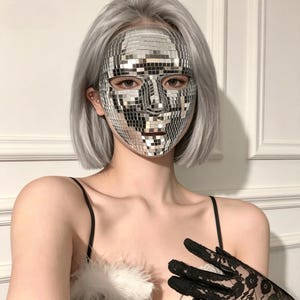 Può includere: Una maschera che copre l'intero volto, rivestita di piccole tessere a specchio argentate e riflettenti. La maschera presenta aperture per occhi, naso e bocca, creando un effetto mosaico che riflette la luce, indossata su un volto con capelli grigio chiaro.