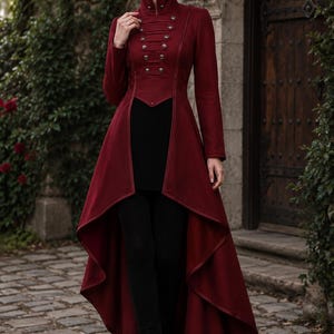 May include: A deep red high-low coat featuring a structured military-style bodice with a high collar, long sleeves, and multiple rows of metallic buttons. The garment has a fitted waist and a dramatic, flowing skirt that is shorter in the front.