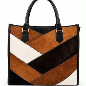May include: A tote bag featuring a geometric patchwork design made of hair-on hide panels in shades of brown, black, and white. The bag has two black leather handles attached with silver-toned metal hardware and black leather trim along the edges.