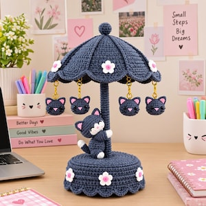 May include: A dark blue crocheted carousel ornament featuring a central pole with a small cat figure attached, a scalloped canopy decorated with white flowers, and four dangling cat head charms suspended by gold-toned metal clasps.