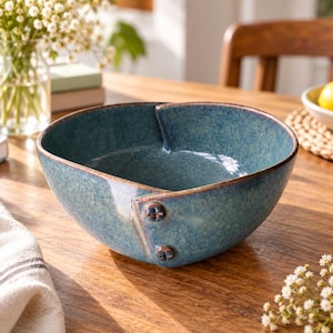 Op de afbeelding: Een diepe, ronde keramische kom met een gespikkeld blauw en groenblauw glazuur. De kom heeft een uniek overlappend ontwerp aan één kant, vastgezet met twee kleine, donkere, cirkelvormige knopachtige accenten in de structuur.