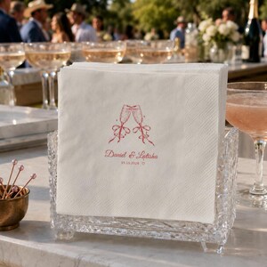 May include: A stack of white paper napkins held in a clear, textured glass holder. The napkins feature a red printed graphic of two clinking champagne flutes with ribbons, the names Daniel & Latisha, and the date 05.15.2026 with a small heart.