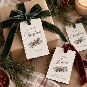Könnte beinhalten: Rechteckige, cremefarbene Geschenkanhänger aus Papier mit Büttenrand, bedruckt mit dem Text Merry Christmas to Emma oder With Love from the Bennett Family, verziert mit grünen Tannennadeln und roten Beeren, befestigt mit dunkelgrünen oder weinroten Samtbändern.