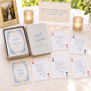 May include: A set of custom playing cards in a silver metal tin, featuring blue and gold floral borders with the text 'The Wedding of Sarah & Charlie December 19, 2026'. Several cards are displayed with handwritten messages from guests.