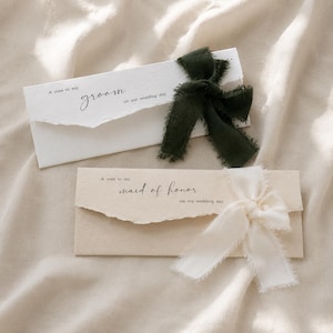 May include: Two rectangular paper envelopes with deckled edges. The top white envelope features a dark green frayed ribbon bow and black text reading A note to my groom on our wedding day. The bottom beige envelope has a cream frayed ribbon bow.