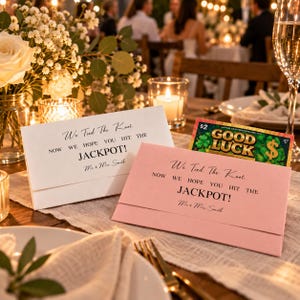 Peut inclure: Deux enveloppes rectangulaires en papier cartonné, une blanche et une rose pâle, imprimées avec le texte noir We Tied The Knot, NOW WE HOPE YOU HIT THE JACKPOT!, et Mr. & Mrs. Smith. Un billet de loterie Good Luck est inséré dans l'enveloppe rose.