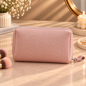 Puede incluir: Un neceser rectangular de color rosa empolvado con textura granulada y cierre de cremallera plateado. El neceser cuenta con un tirador de cremallera a juego de aspecto similar a la piel, sujeto al herraje metálico en el lado derecho.