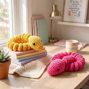Puede incluir: Dos serpientes de peluche enroscadas, una amarilla y otra rosa brillante, con una superficie de ganchillo texturizada y ojos de plástico oscuro. La serpiente amarilla descansa sobre una pila de cuadernos, mientras la rosa está en el escritorio.