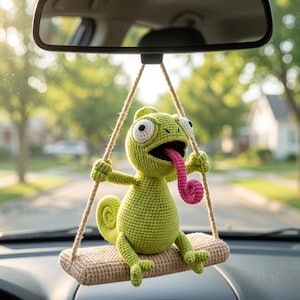 Puede incluir: Un juguete de camaleón tejido a ganchillo en color verde claro con grandes ojos blancos y una larga lengua rosa, sentado en un columpio rectangular tejido en color beige suspendido por dos cuerdas, para colgar del espejo retrovisor.