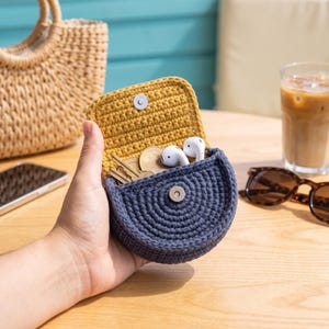 May include: A small, semi-circular crochet pouch featuring a dark gray exterior and a mustard yellow interior flap. The pouch is secured with a silver-toned metal snap button and contains white wireless earbuds and several metal keys inside.