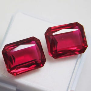 May include: Two rectangular, faceted, deep red gemstones with sharp corners and a high-gloss finish. The stones exhibit a vibrant, saturated crimson hue with clear internal reflections and light refraction across their polished surfaces.