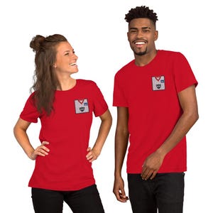 May include: A bright red short-sleeved t-shirt featuring a small rectangular graphic on the left chest. The graphic depicts a white and grey striped sweater vest with a red V-neck collar, a small blue square, and a black shield logo with the text HSV.