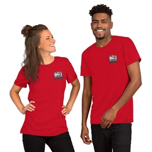 Könnte beinhalten: Ein leuchtend rotes Kurzarm-T-Shirt mit einer kleinen rechteckigen Grafik auf der linken Brust. Die Grafik zeigt ein stilisiertes Trikotdesign mit horizontalen Streifen in Grau, Weiß und Dunkelrot sowie dem in Schwarz gedruckten Wort BAYERN.