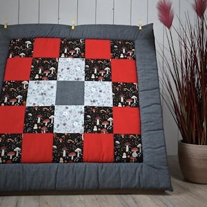 Könnte beinhalten: Eine quadratische Patchwork-Decke mit einem zentralen grauen Block, umgeben von weißen Blumenquadraten und schwarzen Quadraten mit Pilzmotiv. Die Decke enthält einfarbige rote Quadrate und ist von einem breiten, dunkelgrauen Rand mit Nähten umgeben.