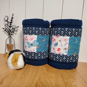 Könnte beinhalten: Zwei zylindrische Stofftunnel mit einer marineblauen Fleece-Außenseite und einem Mittelstreifen aus gemustertem Baumwollstoff mit Ankern, Schmetterlingen und Blumenmotiven in Weiß, Blau und Rosa, daneben ein kleines Meerschweinchen-Stofftier.