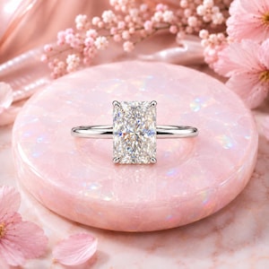 May include: A silver-toned ring featuring a large, rectangular radiant-cut centre stone with multiple facets. The stone is held in place by four prongs on a thin, polished metal band. The design is centred and symmetrical, showcasing clear, reflective surfaces.