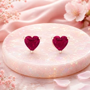 Puede incluir: Un par de pendientes de botón en forma de corazón con piedras facetadas de color rojo intenso montadas en un marco metálico. Cada pendiente está asegurado con una pequeña garra en la punta inferior, mostrando un tono carmesí vibrante y brillante.