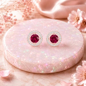 May include: A pair of round stud earrings featuring a vibrant deep red circular gemstone at the center, surrounded by a halo of small, clear, sparkling white stones set in a polished rose gold metal frame with a classic post-back design.