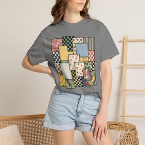 Peut inclure: Un t-shirt gris anthracite à manches courtes avec un imprimé graphique central représentant un motif de patchwork. Les pièces rectangulaires présentent divers motifs, notamment des pois, des carreaux, des fleurs, des rayures et des étoiles.